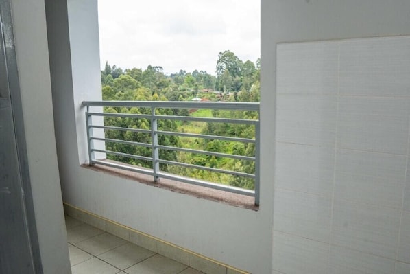 Un balcon avec une rambarde en métal, une fenêtre et une vue sur la verdure et les bâtiments.