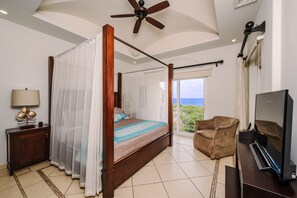 Master bedroom foot-of-bed view showing four-poster king, louvered hardwood closets, TV, and hallway to en-suite. - Ocean View Condo with Huge Lagoon Pool Across from the Beach (Tamarindo)
