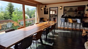 Uma sala de jantar com uma mesa de madeira comprida, cadeiras e vista para áreas verdes e montanhas através de grandes janelas.