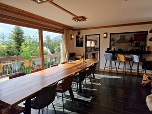Ein Essbereich mit einem langen Holztisch, Stühlen und Blick auf Grünflächen und Berge durch große Fenster.