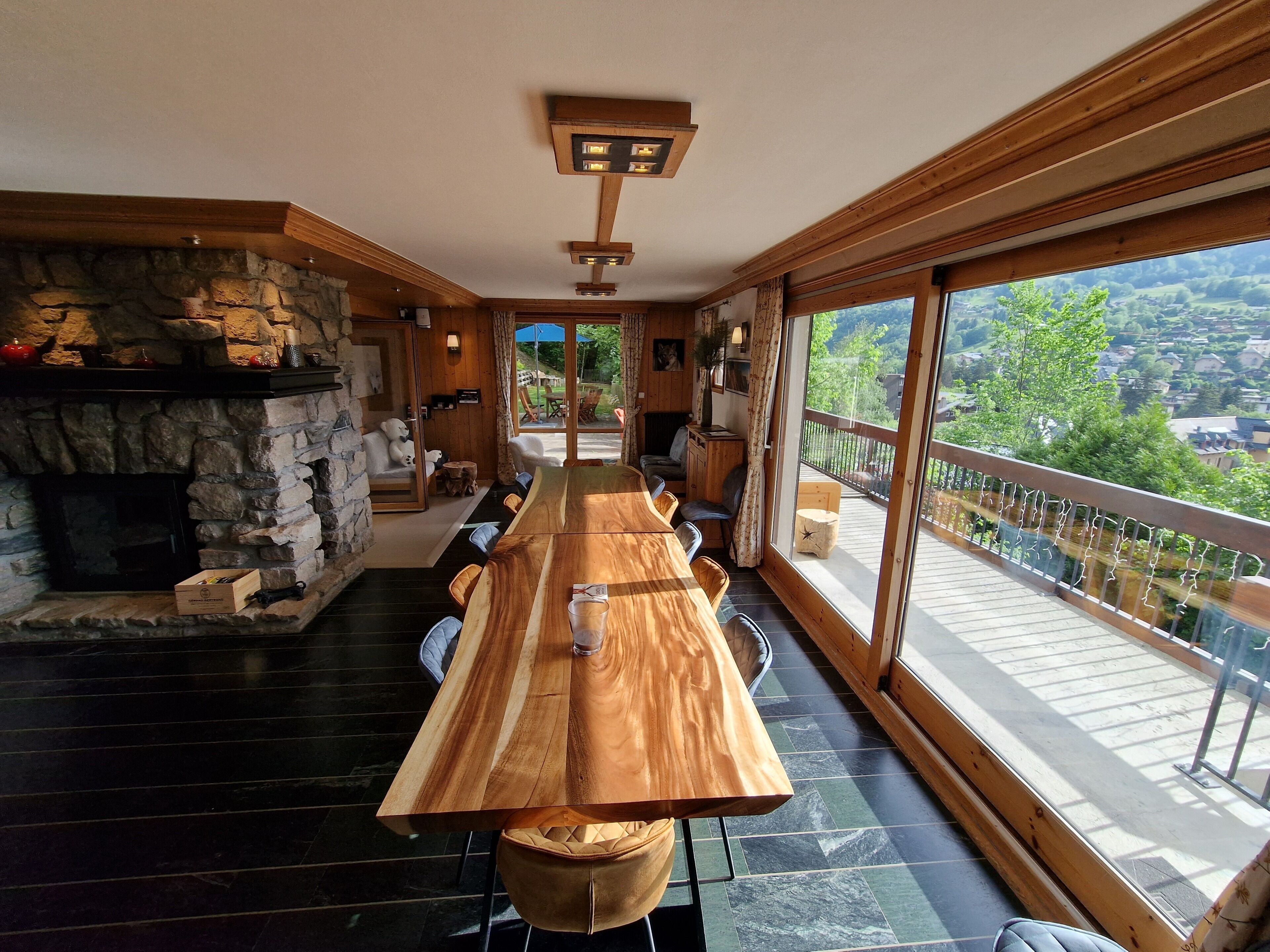 Une longue table à manger en bois, entourée de chaises, une cheminée en pierre et de grandes fenêtres donnant sur l’extérieur.