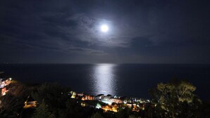 Uma cidade costeira à noite com uma lua cheia proeminente, edifícios iluminados e vista para o mar.