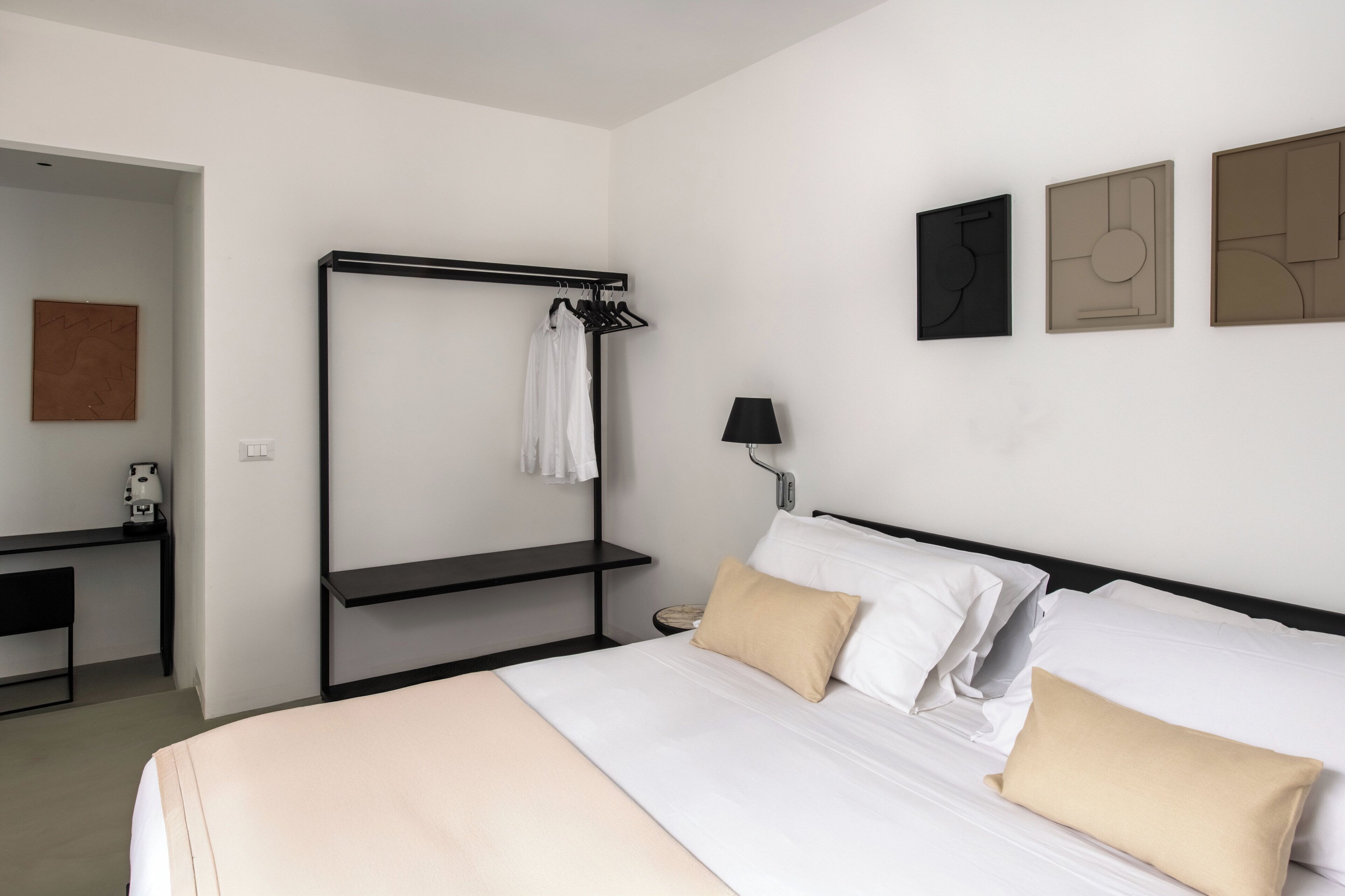 Una habitación de hotel con cama, mesita de noche, lámpara, perchero y una obra de arte en la pared.