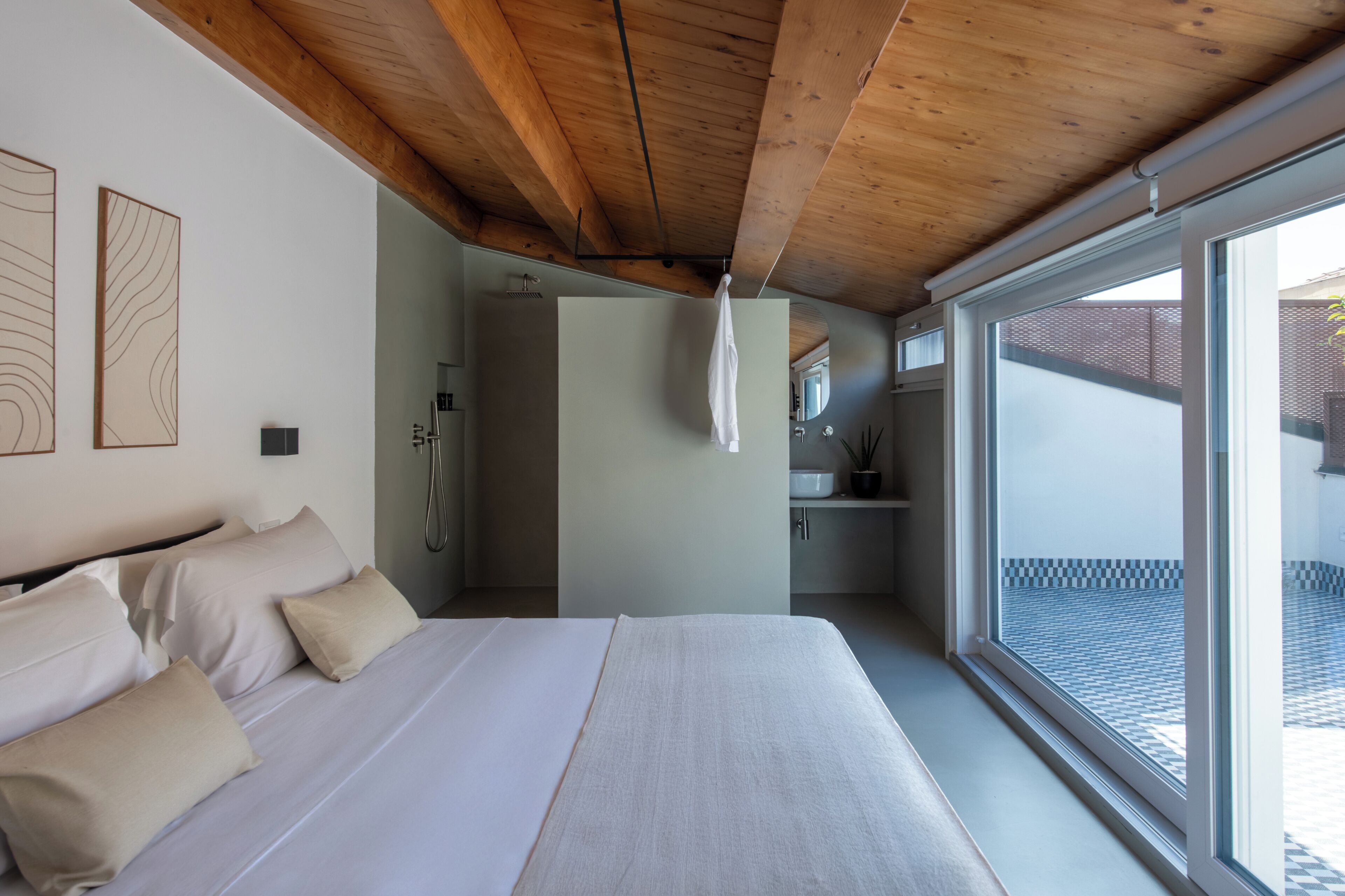 A bedroom with a large bed, wooden ceiling beams, a bathroom with a shower, and a view through a large window.