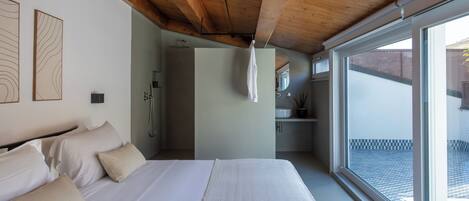 A bedroom with a large bed, wooden ceiling beams, a bathroom with a shower, and a view through a large window.