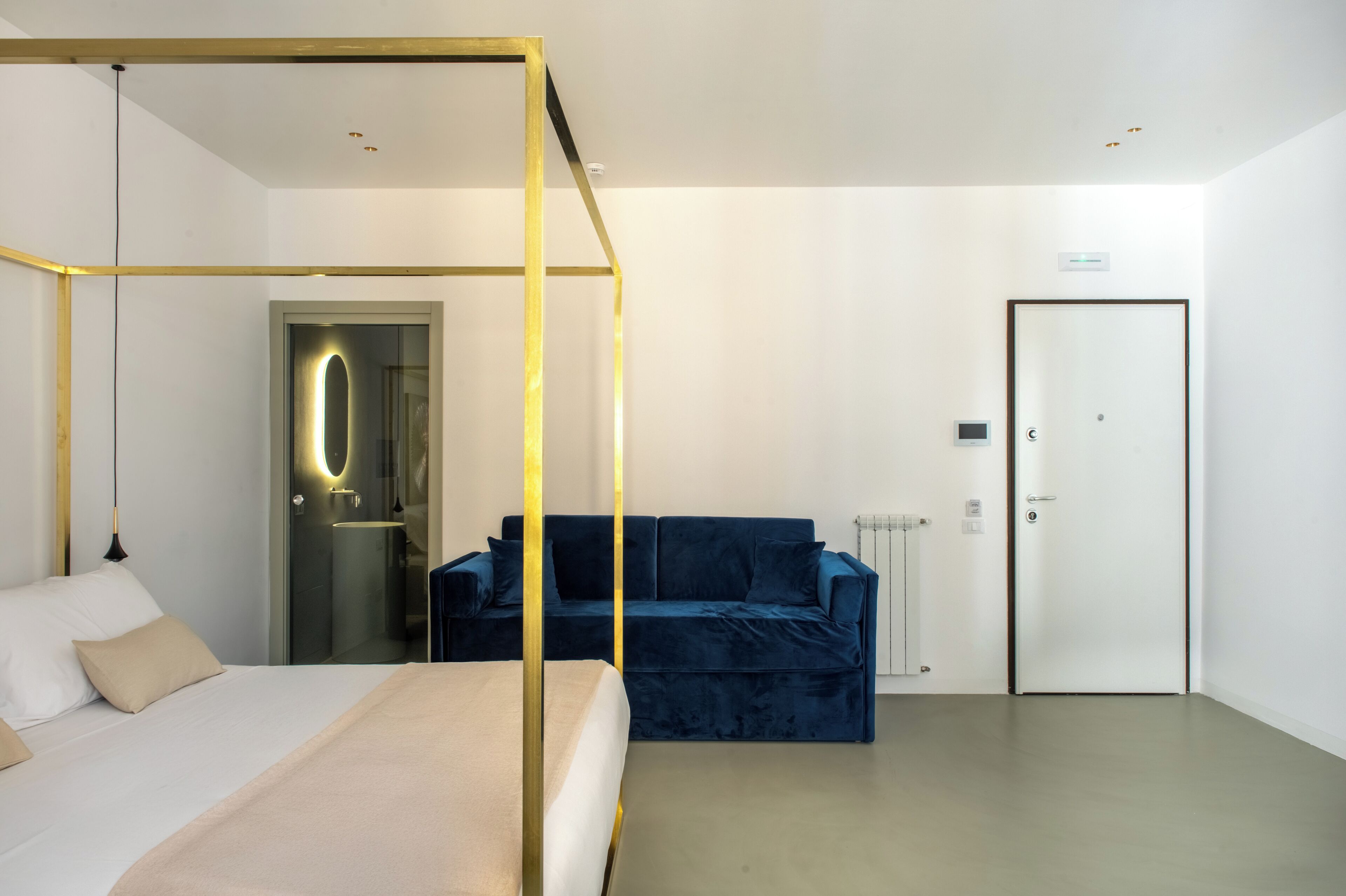 A modern hotel room with a four-poster bed, a blue sofa, a white door, and a glass-enclosed shower area.