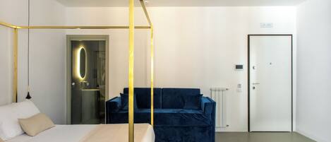 A modern hotel room with a four-poster bed, a blue sofa, a white door, and a glass-enclosed shower area.