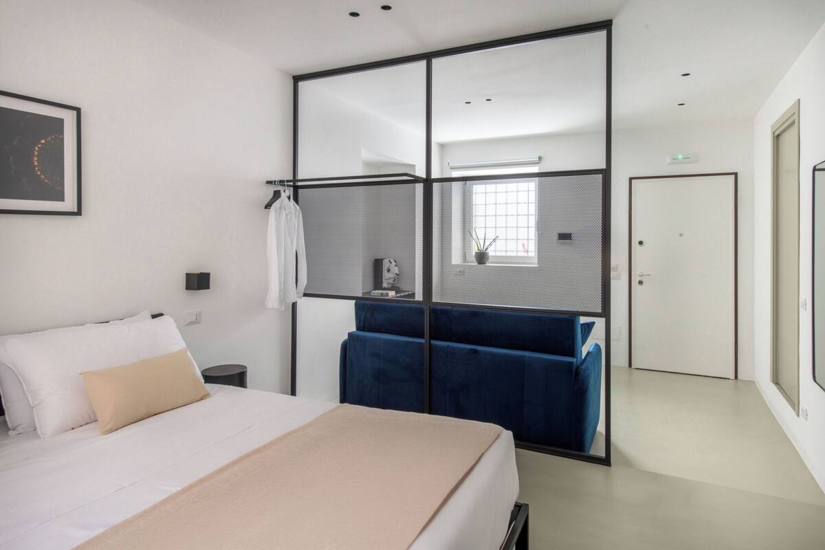 Una habitación de hotel moderna con cama, un banco tapizado en azul, una mesita con planta en maceta y una puerta corredera.