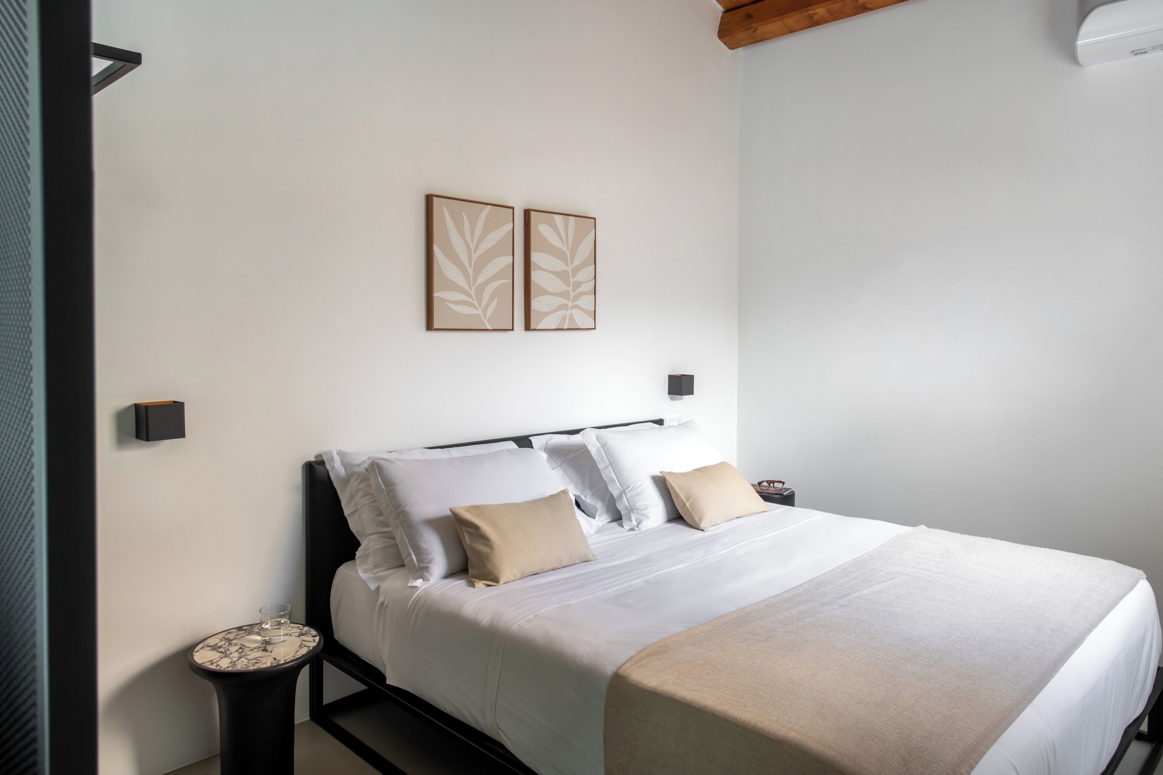 A neatly made bed with white linens and beige pillows, a small round table with a glass and a book, and two wall-mounted artworks.