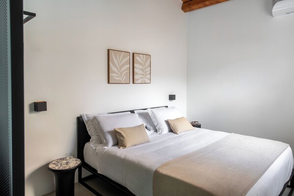 A neatly made bed with white linens and beige pillows, a small round table with a glass and a book, and two wall-mounted artworks. - Palazzo Achates (Palermo)
