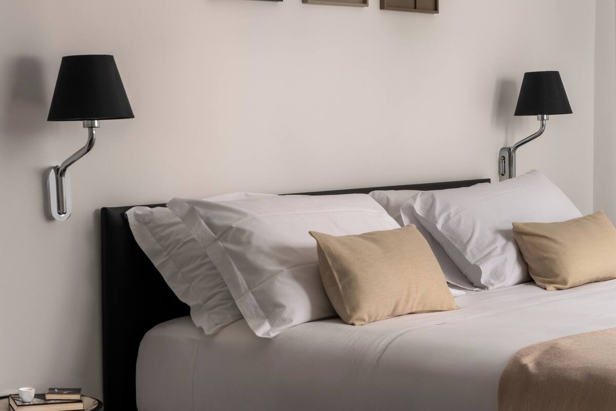 Una cama bien hecha con almohadas blancas y beige, dos lámparas de pared y una mesita con libros y una taza.