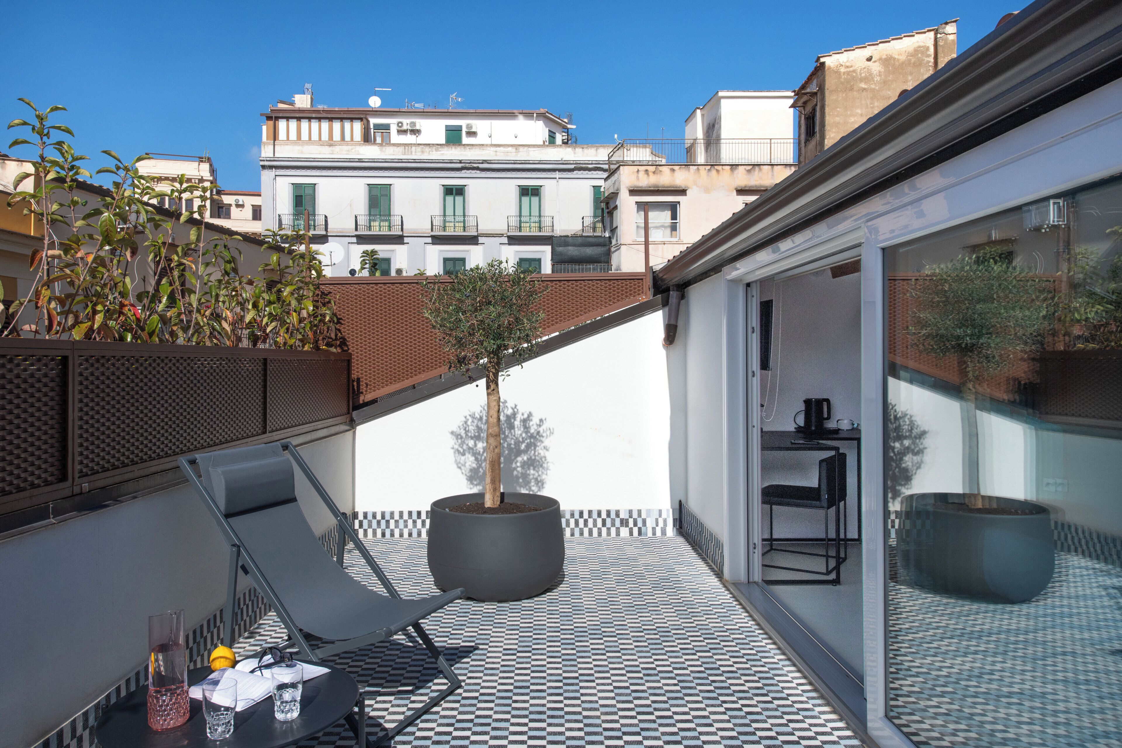 Una terraza en la azotea con piso a cuadros, un árbol en maceta, una silla de lounge y un área de comedor con paredes de vidrio.