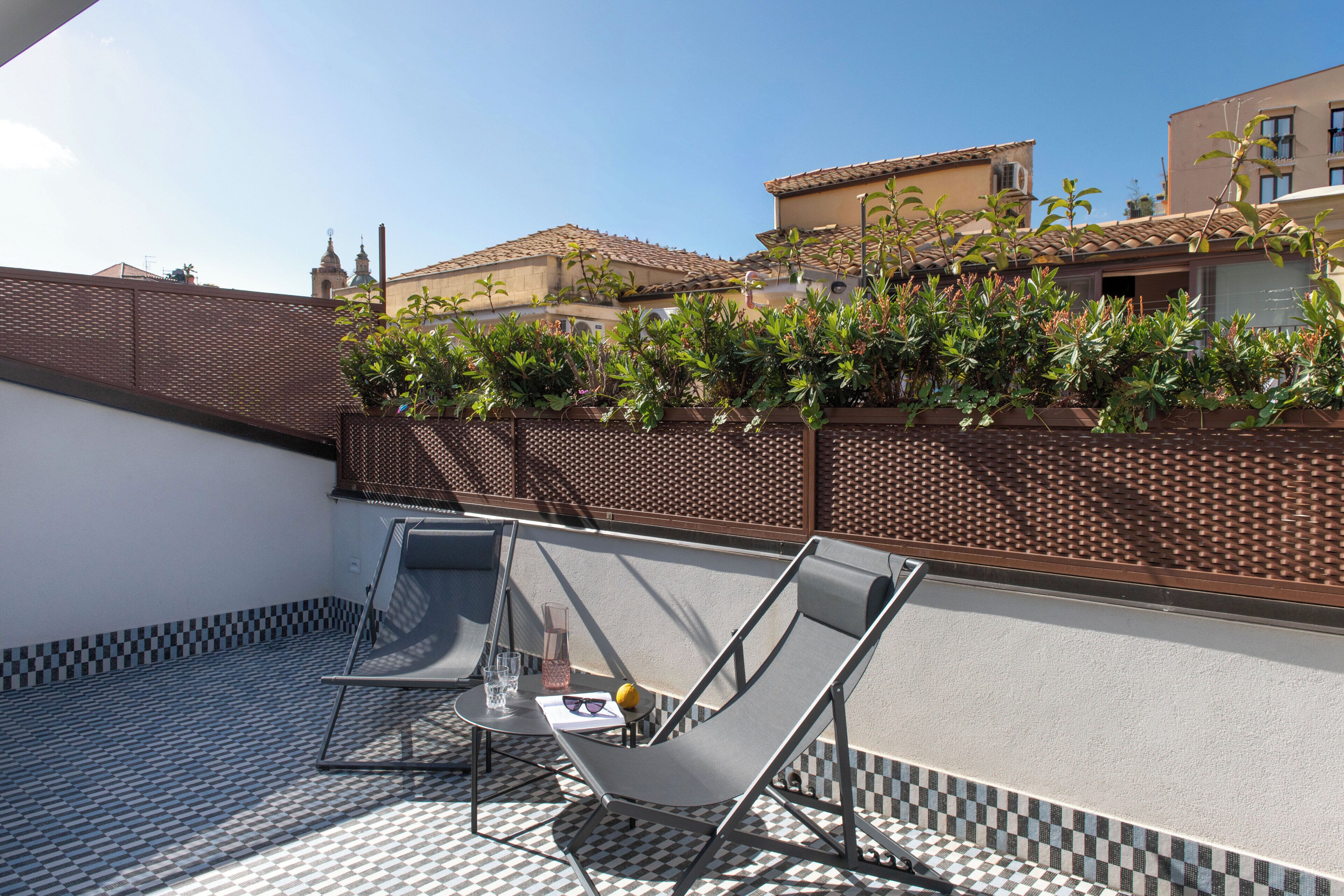 Eine Dachterrasse mit Schachbrettboden, einem Glastisch, einem Metallstuhl und Blick auf Gebäude im Hintergrund.