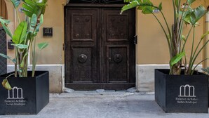 A building facade with a large arched entrance, a balcony with metal railings, and two potted plants with broad leaves. - Palazzo Achates (Palermo)