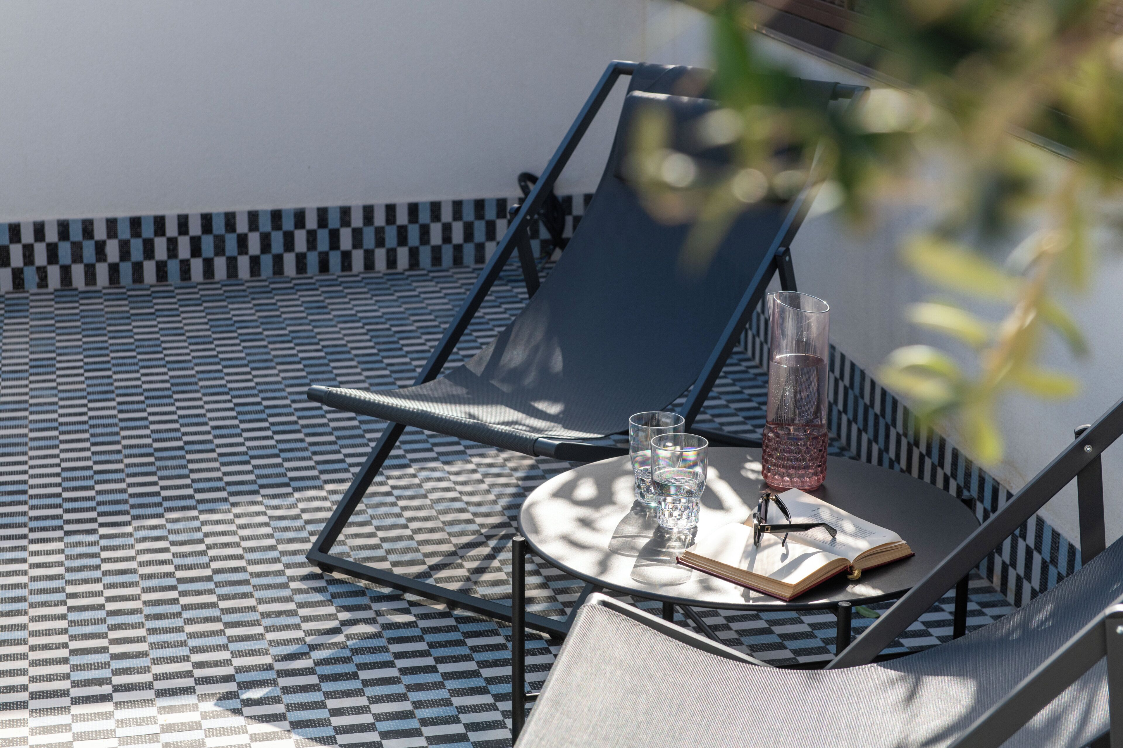 A sunlit outdoor seating area with a checkered floor, two lounge chairs, a round table with a book, glasses, and a water bottle.