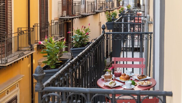 A balcony with a table set for a meal, potted plants, and a railing. - Palazzo Achates (Palermo)
