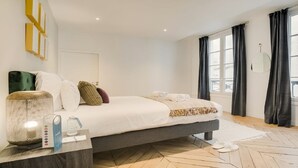 A modern bedroom with a bed, bedside table, lamp, and large windows with curtains. - Bonne Nouvelle Beauty | 200m² of Parisian Elegance Near Opéra (Paris)