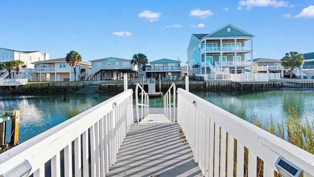 A waterfront property with a wooden dock leading to a pier, surrounded by buildings with balconies. - Fabulous 4-bedroom channel home with private dock in Cherry Grove Beach. (North Myrtle Beach)