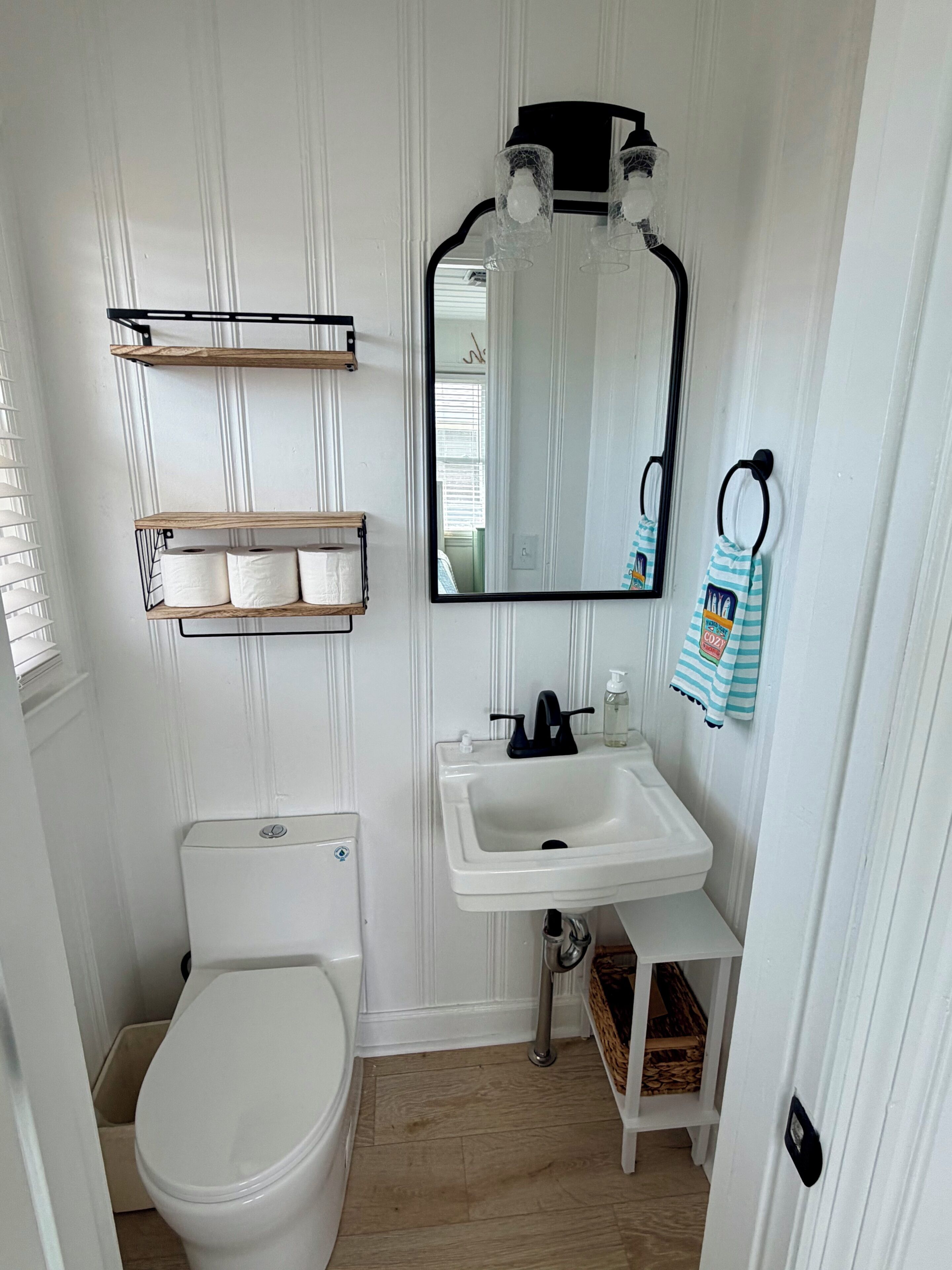 Et lille badeværelse med toilet, håndvask, spejl og hylder.