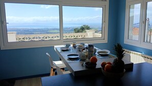 A dining area with a table set for two, a bowl of fruit, and a view of the landscape through large windows. - Casa Peroscalou Roses – Modern Villa with Panoramic Views, 3 Bedrooms, Near Costa Brava Beaches (El Mas Fumats)