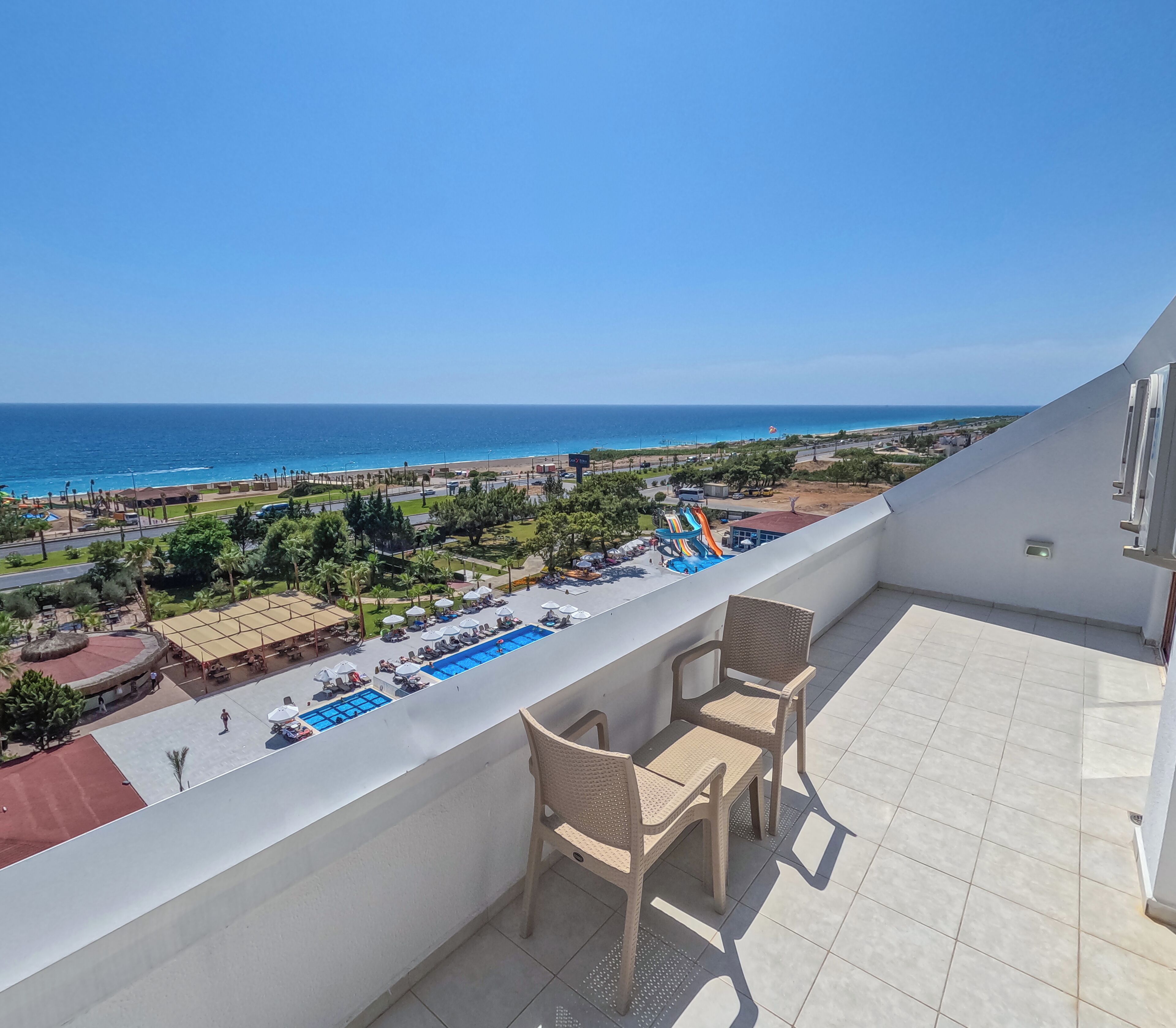 Un balcón con vistas a una piscina y una playa, sillas de mimbre y cielo despejado.