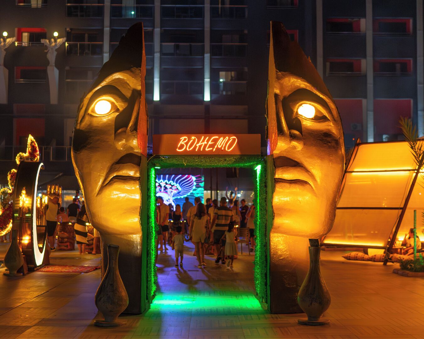 Una entrada amplia e iluminada con un letrero de neón que dice 'BOHEMO' y un camino que conduce a un edificio.