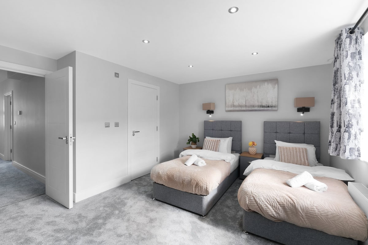 A modern bedroom with two beds, a grey headboard, a large abstract painting, and a door leading to another room.