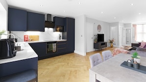 A modern kitchen with dark cabinetry, a dining area with a table and chairs, and a living room with a sofa and TV. - 4BDR 3BA Family Home Garden in Isleworth (Isleworth)