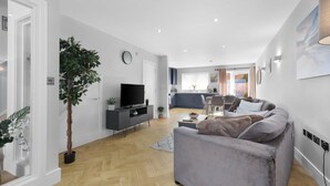 A modern living room with a grey sofa, a TV on a black stand, a potted plant, and a wooden floor. - 4BDR 3BA Family Home Garden in Isleworth (Isleworth)