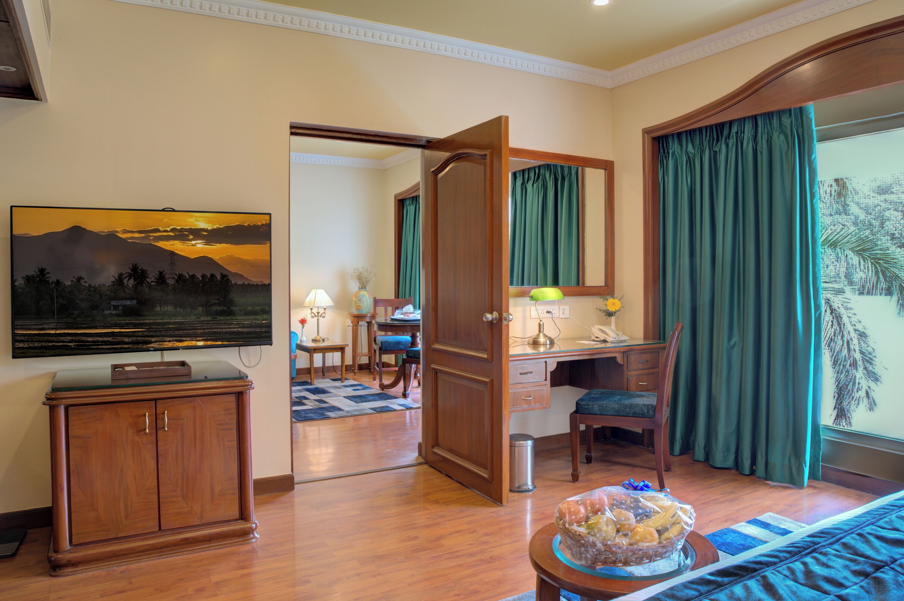 A hotel room with a wooden desk, a television, a dining area, and a large window with curtains.