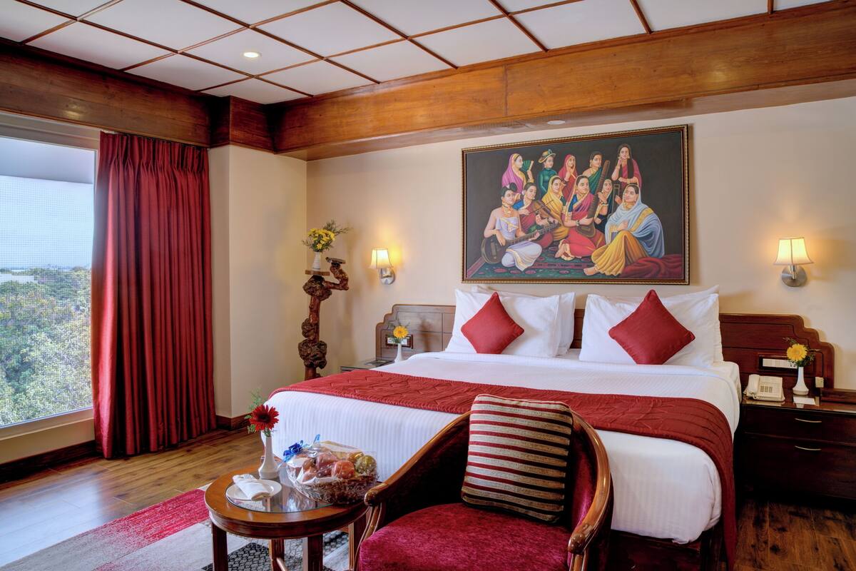 Habitación de hotel con una cama grande, cabecera de madera, un cuadro en la pared, una mesita de noche con lámpara y vistas al exterior.