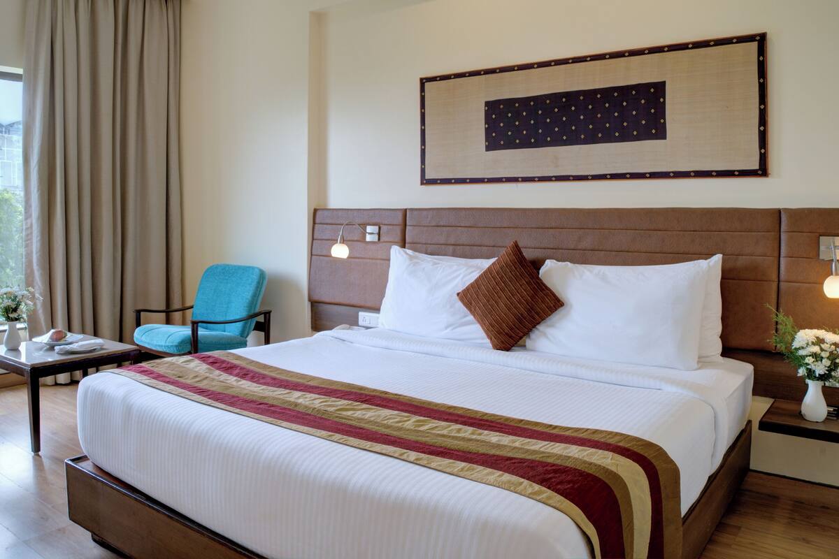 Una habitación de hotel con una cama grande, un sillón azul, una mesita de noche con un jarrón de flores y una ventana con cortinas.