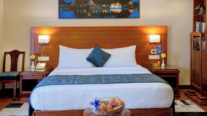 A hotel room with a large bed, wooden headboard, two bedside lamps, a nightstand, a small table with a decorative item, and a painting above the bed. - Hotel Tamilnadu Pandiyan Madurai (Madurai)