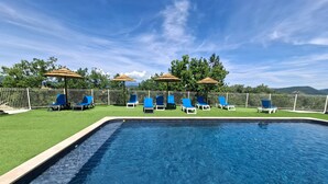 A swimming pool with blue lounge chairs and umbrellas, surrounded by a white fence and green grass.