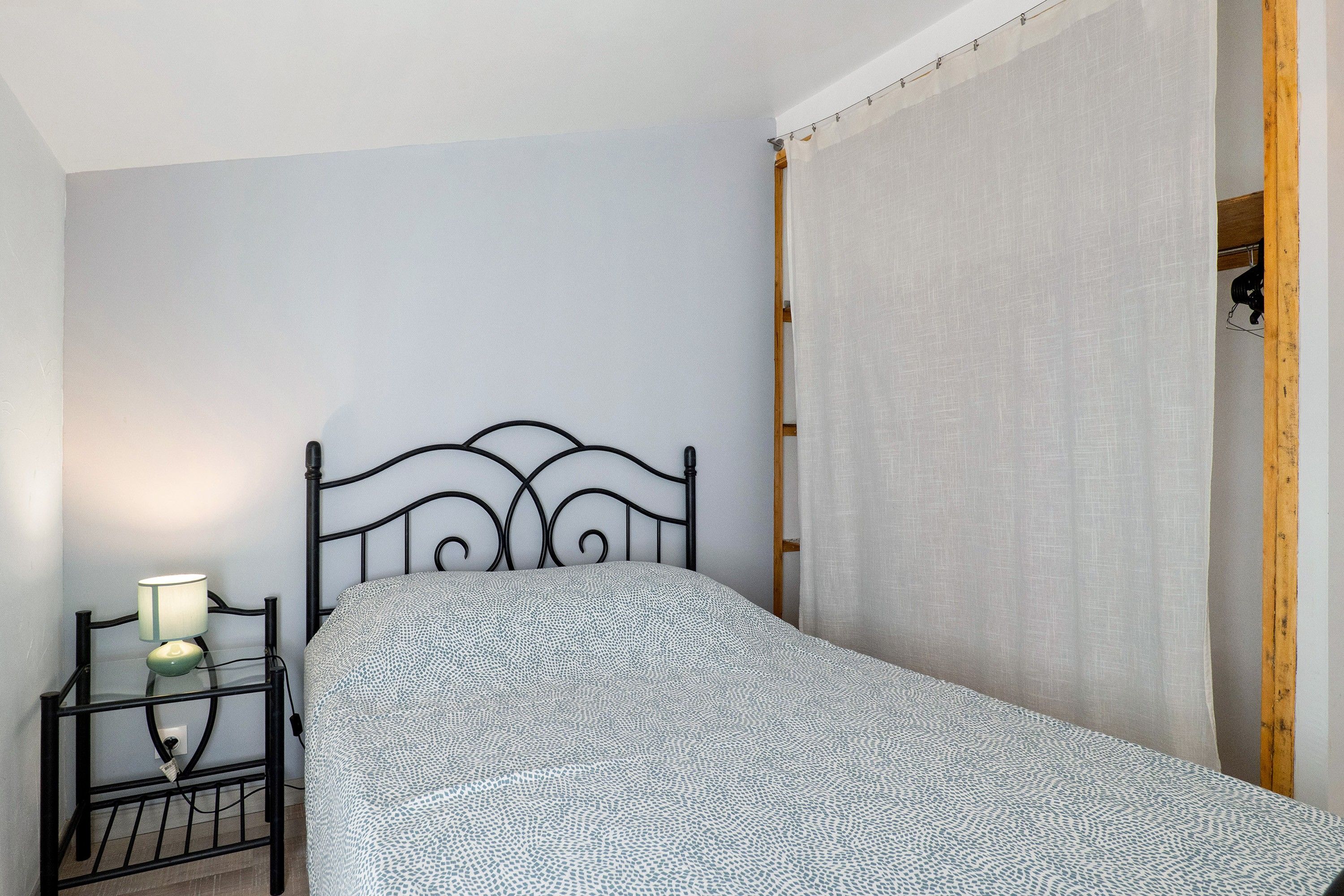 Una cama individual con colcha estampada en una habitación con paredes blancas, una mesita de noche con lámpara y una puerta de madera.