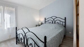 Uma cama de solteiro com estrutura de metal em quarto com parede azul claro, janela com cortinas e mesa de cabeceira com luminária.