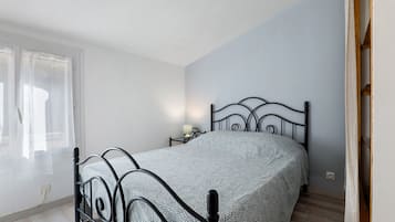 Ein Einzelbett mit Metallgestell in einem Zimmer mit hellblauer Wand, einem Fenster mit Vorhängen und einem Nachttisch mit Lampe.