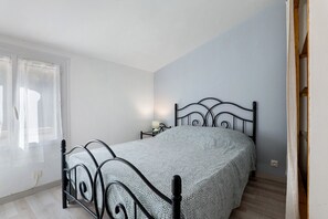 Een eenpersoonsbed met metalen frame in een kamer met een lichtblauwe muur, een raam met gordijnen en een nachtkastje met een lamp.