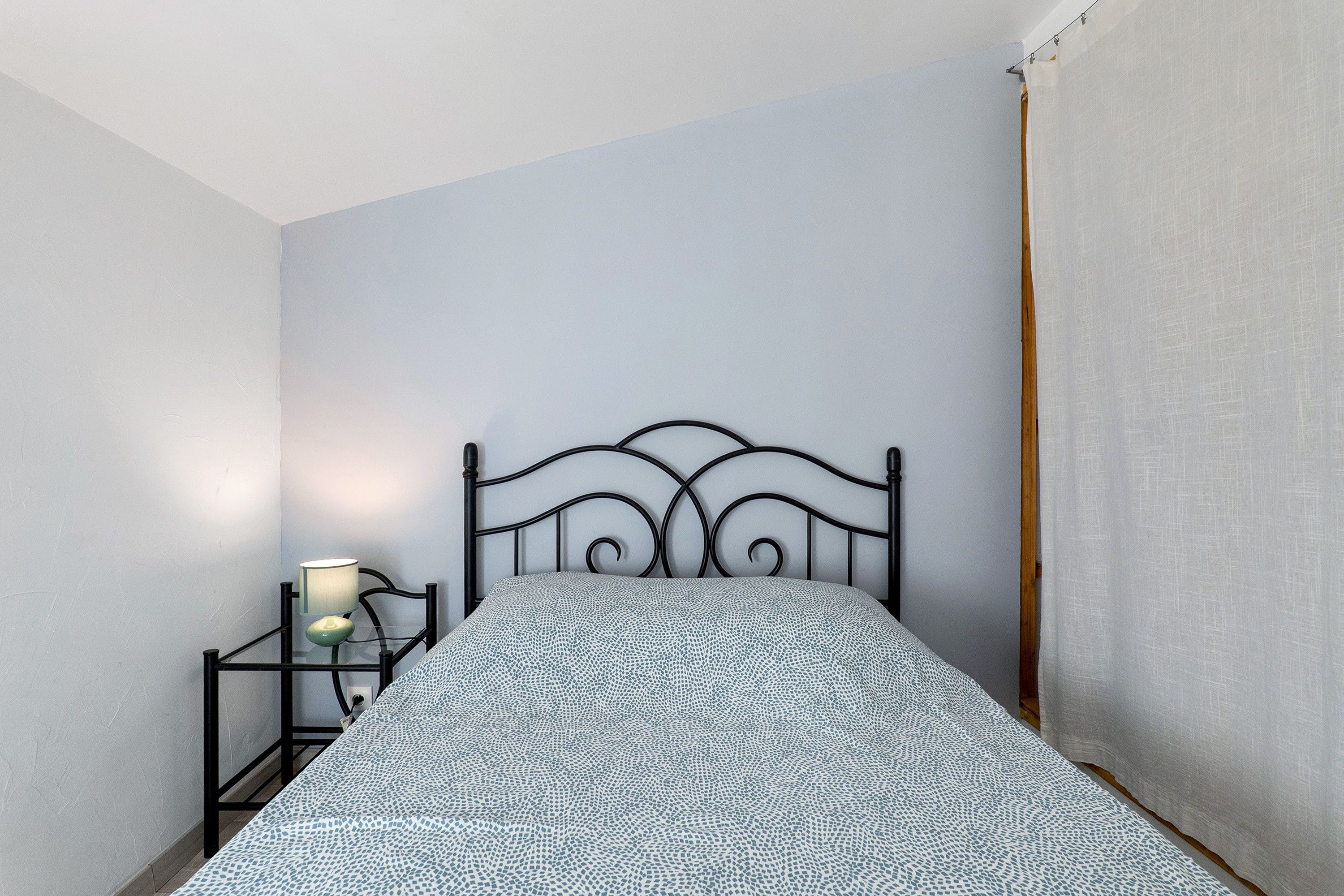 Een enkel bed met een metalen frame en een beddegoed met patroon in een kamer met lichtgekleurde wanden en een gordijn aan de zijkant.