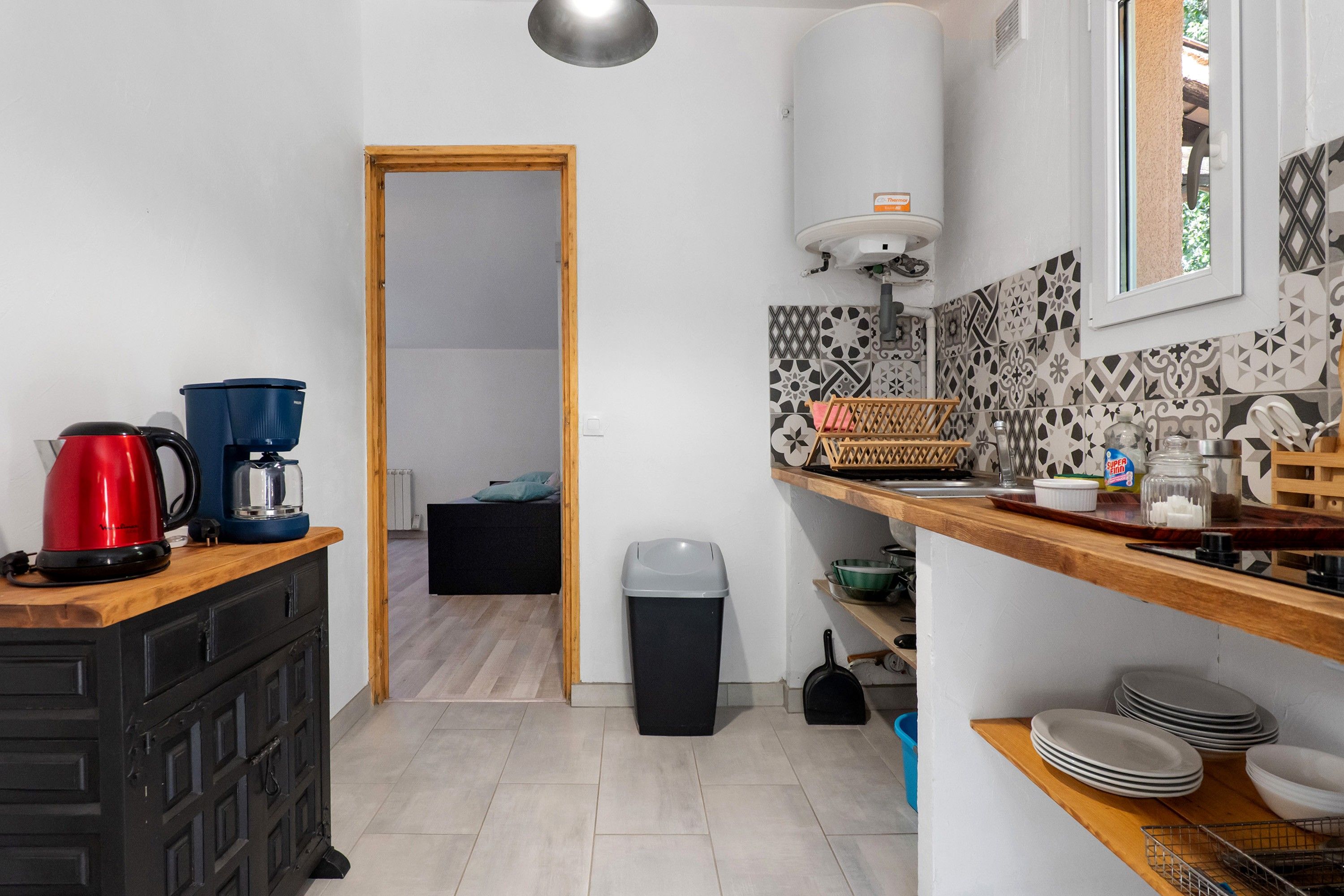 Une petite cuisine équipée d’une machine à café, d’une bouilloire et d’une poubelle. La pièce adjacente contient un lit et un radiateur est fixé au mur.