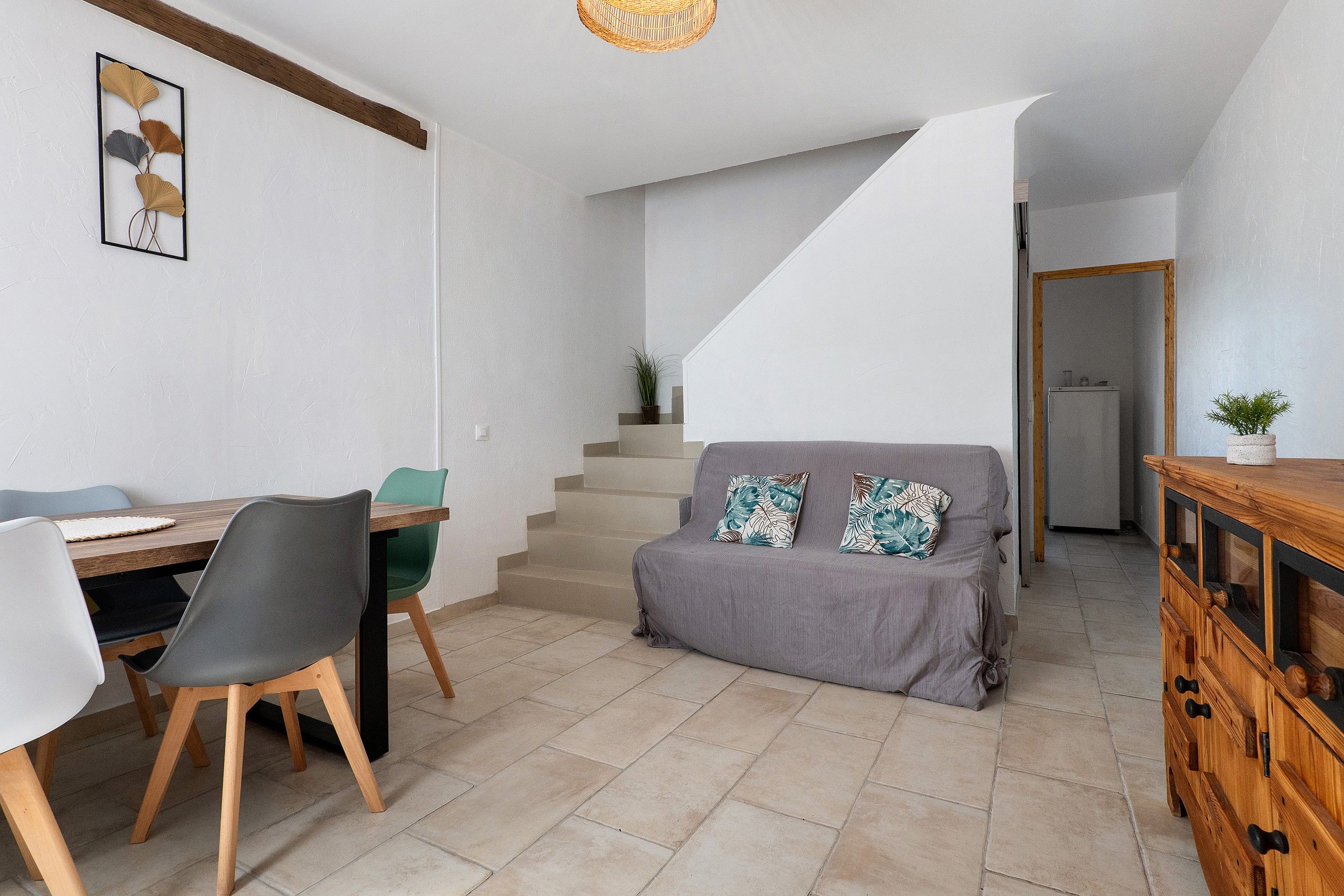 A dining area with a wooden table and chairs, a staircase leading to an upper level, and a grey sofa with patterned cushions.