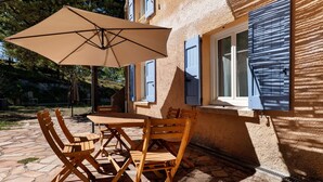 Eine Terrassengruppe im Freien mit Tisch und Stühlen, einem beige gefärbten Sonnenschirm und einem Gebäude mit blauen Fensterläden.