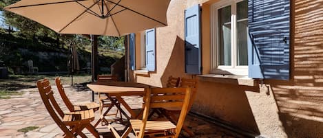 An outdoor patio with a table and chairs, a beige umbrella, and a building with blue shutters.