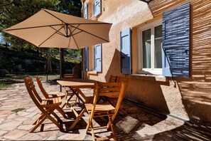 Eine Terrassengruppe im Freien mit Tisch und Stühlen, einem beige gefärbten Sonnenschirm und einem Gebäude mit blauen Fensterläden.