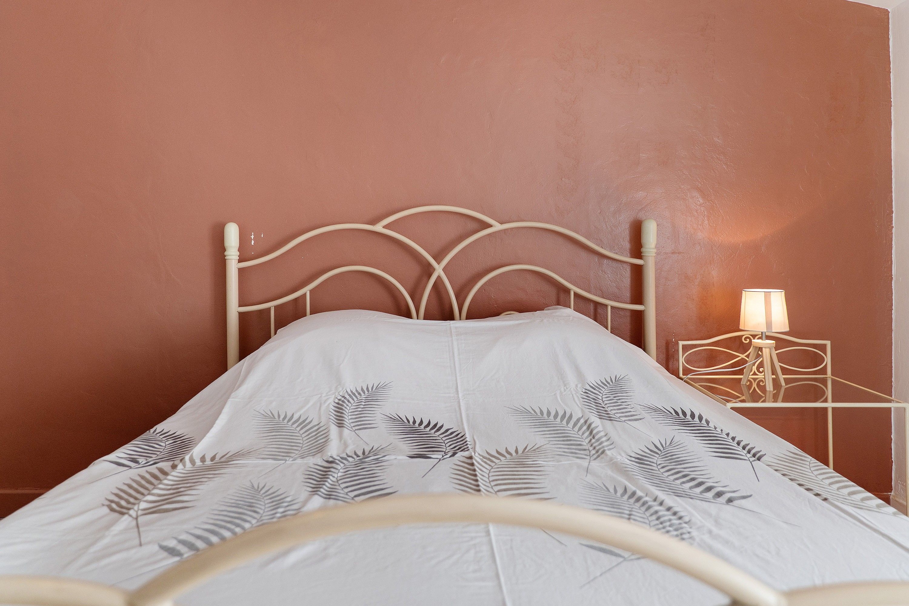 Ein Bett mit weißem Kopfteil und einer geblümten Tagesdecke, ein Nachttisch mit Lampe sowie eine Wand mit strukturierter Oberfläche.