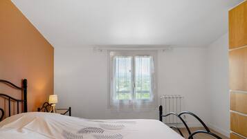 Un dormitorio con cama, mesita de noche, lámpara, ventana con cortinas y radiador.