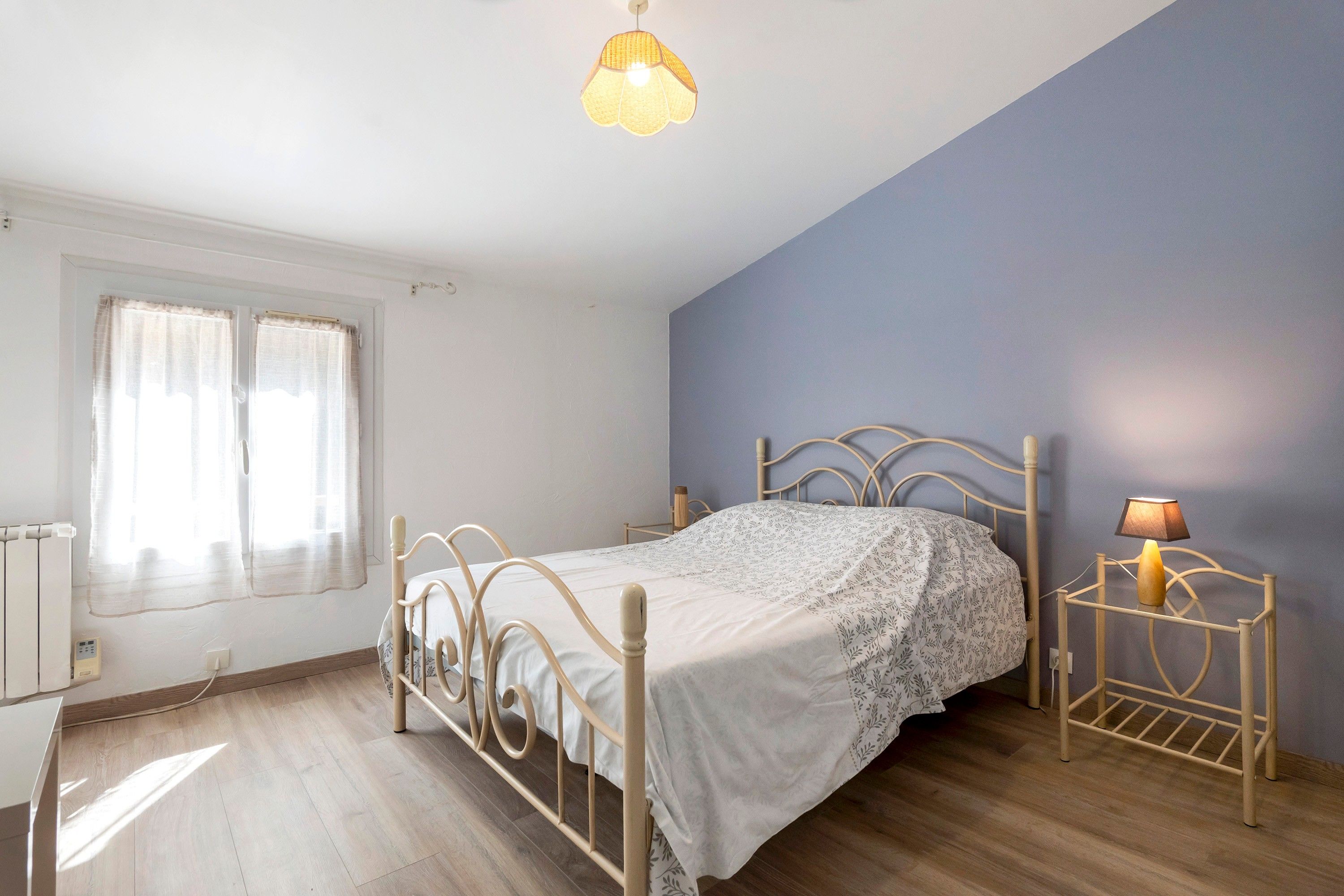 A bedroom with a bed, a bedside table with a lamp, a window with curtains, and a radiator.