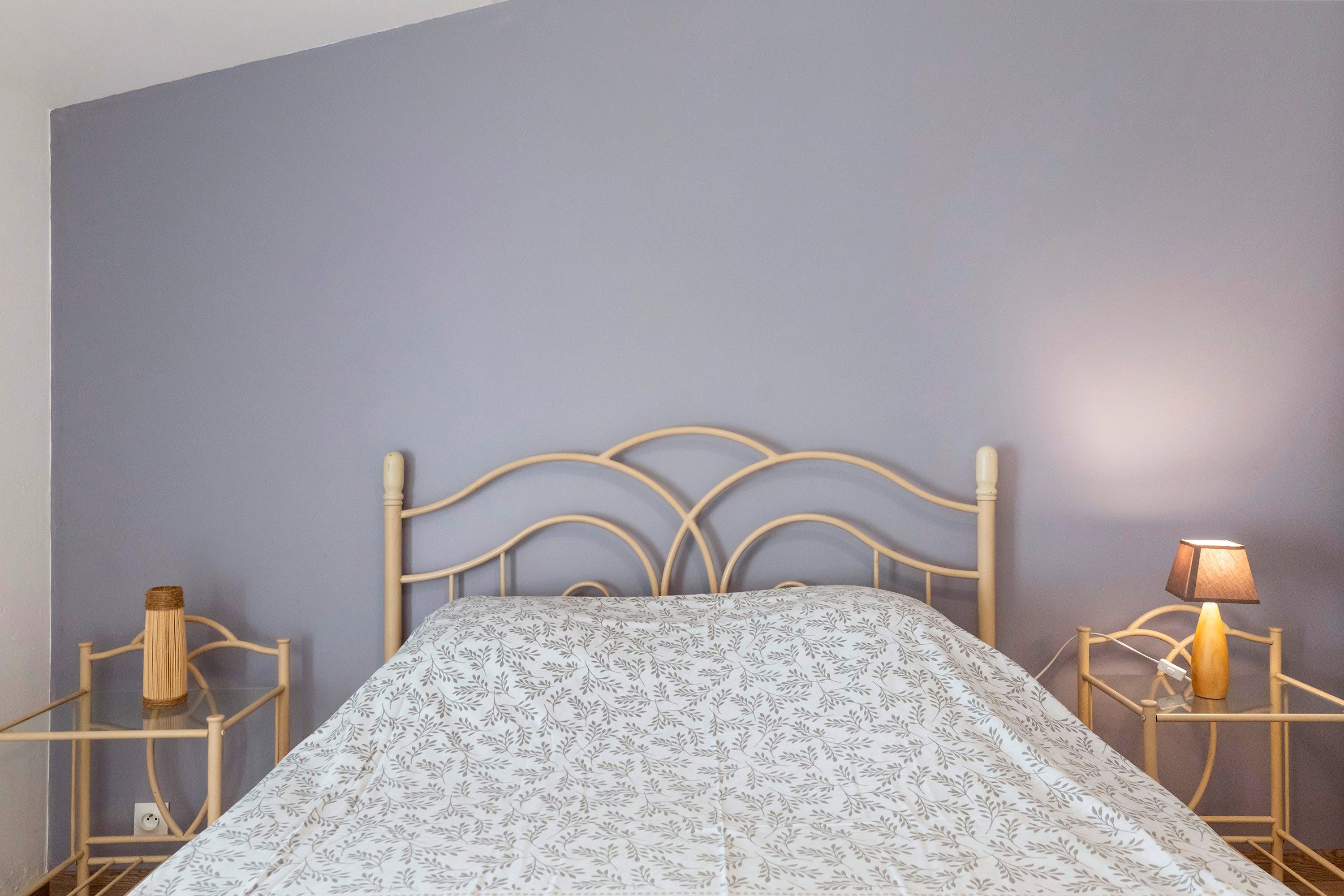 Un llit amb estructura metàl·lica i un edredó estampat, flanquejat per taules de nit amb làmpades, davant d'una paret sense decoració.
