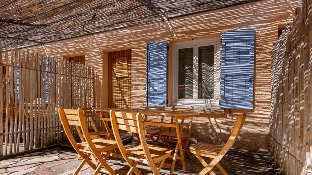 Un espace extérieur aménagé avec des meubles en bois, comprenant une table et des chaises, un toit de chaume et une fenêtre ornée de volets bleus.