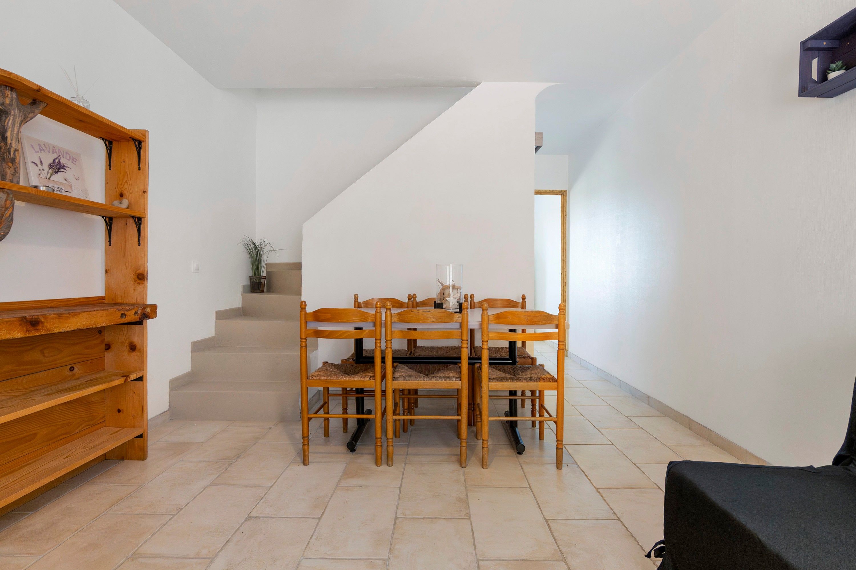 A dining area with a wooden table and chairs, a staircase leading to another level, and a shelf with decorative items.